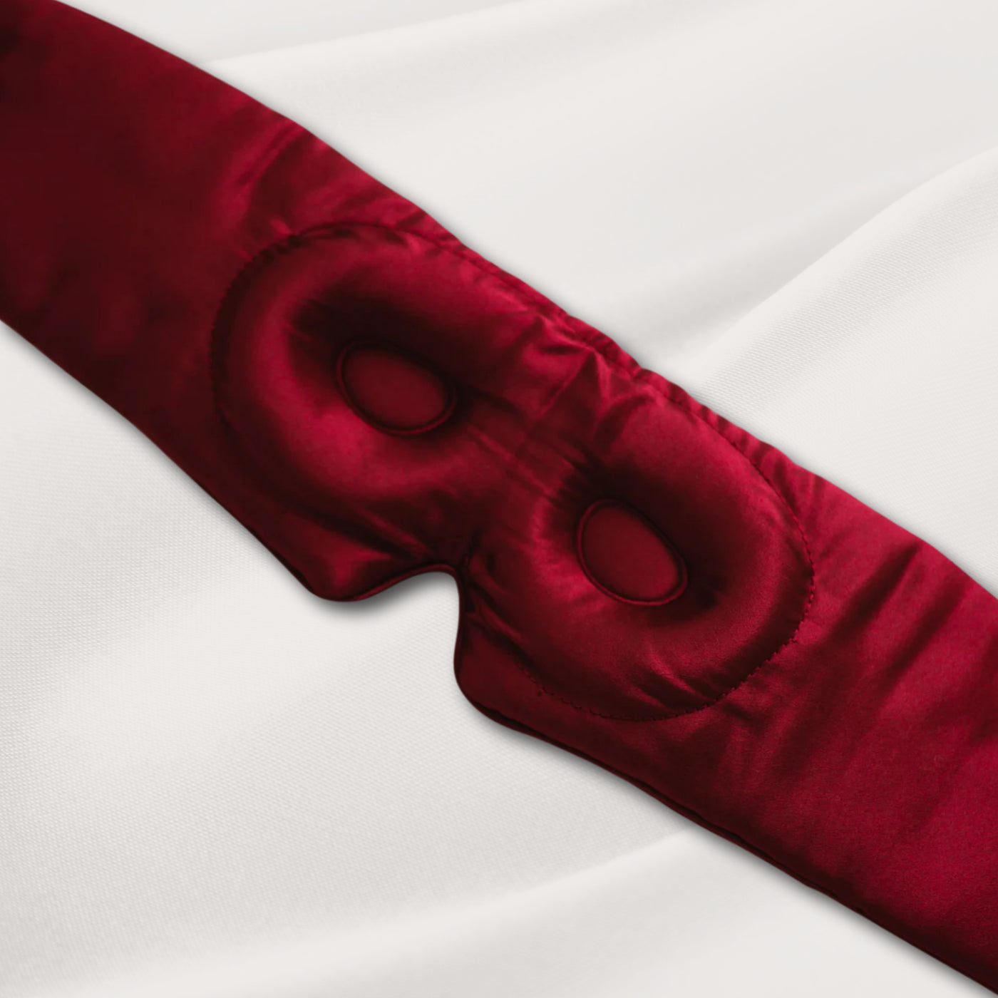 Burgundy LashGuard 3D sleep mask resting on white bedding, angled view of the contoured eye cups