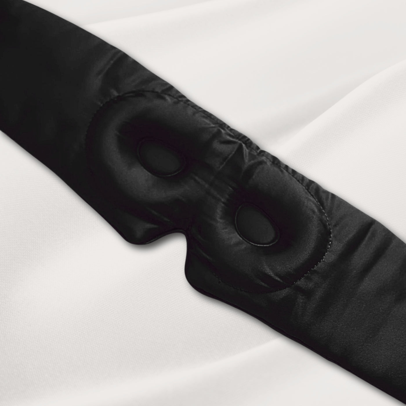 Black LashGuard 3D sleep mask resting on white bedding, angled view of the contoured eye cups